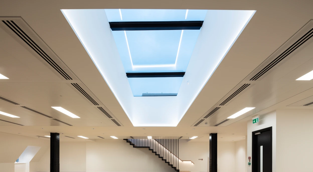 Espacio interior moderno con suelos de madera y una escalera elegante. Hay una claraboya grande en el techo que permite la entrada de luz natural. Las paredes son claras y el ambiente es minimalista.