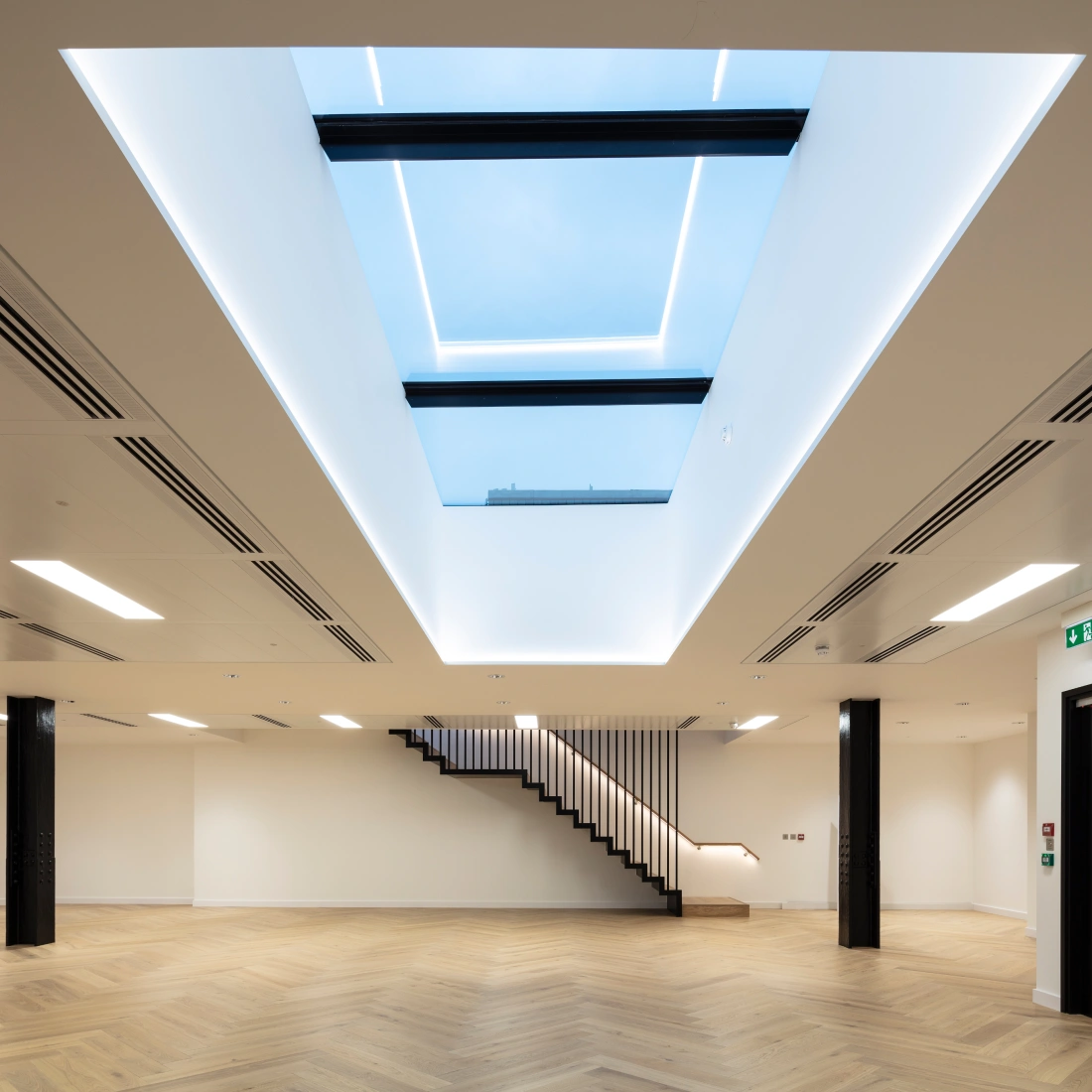 Espacio interior moderno con suelos de madera y una escalera elegante. Hay una claraboya grande en el techo que permite la entrada de luz natural. Las paredes son claras y el ambiente es minimalista.