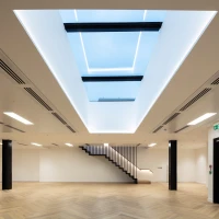 Espacio interior moderno con suelos de madera y una escalera elegante. Hay una claraboya grande en el techo que permite la entrada de luz natural. Las paredes son claras y el ambiente es minimalista.