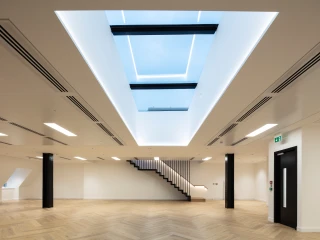 Offener Raum mit hellen Wänden und einem großen Dachfenster. Dezente LED-Beleuchtung und elegante Holztreppe im Hintergrund. Der Boden ist aus Holz mit einem Fischgrätenmuster.