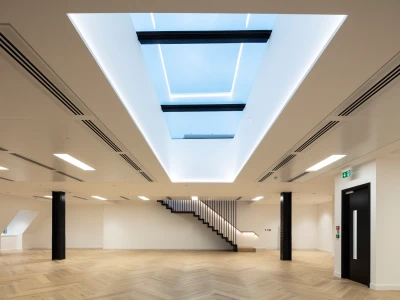 Intérieur moderne avec un plafond lumineux et un puits de lumière. Un escalier en bois blanc mène à un niveau supérieur. Le sol est en parquet clair et les murs sont peints en blanc.