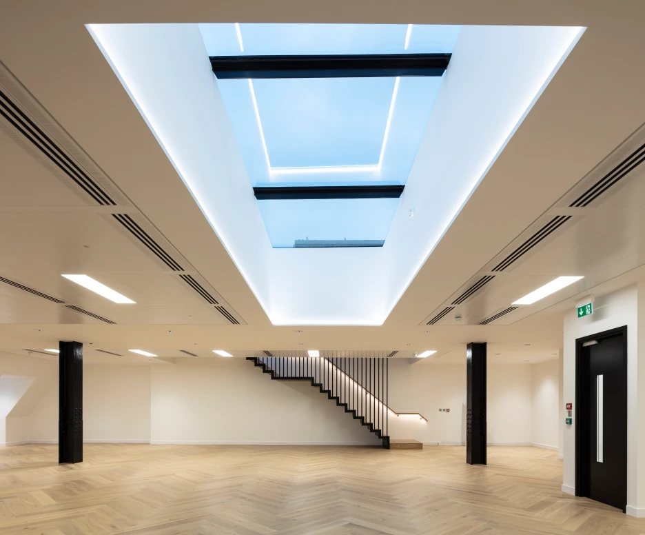 Offener Raum mit hellen Wänden und einem großen Dachfenster. Dezente LED-Beleuchtung und elegante Holztreppe im Hintergrund. Der Boden ist aus Holz mit einem Fischgrätenmuster.