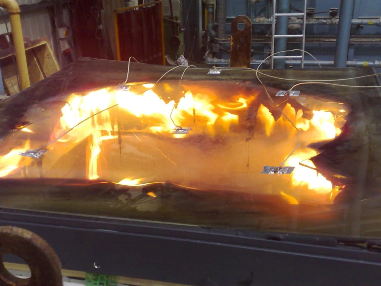 A large, flat surface is shown with flames and smoke on it, suggesting a controlled fire experiment. Metal wires are attached to the surface, indicating monitoring equipment in an industrial or laboratory setting.