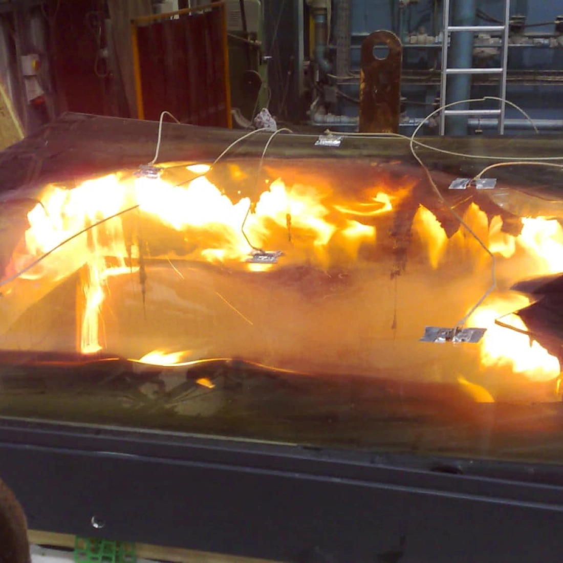A large, flat surface is shown with flames and smoke on it, suggesting a controlled fire experiment. Metal wires are attached to the surface, indicating monitoring equipment in an industrial or laboratory setting.