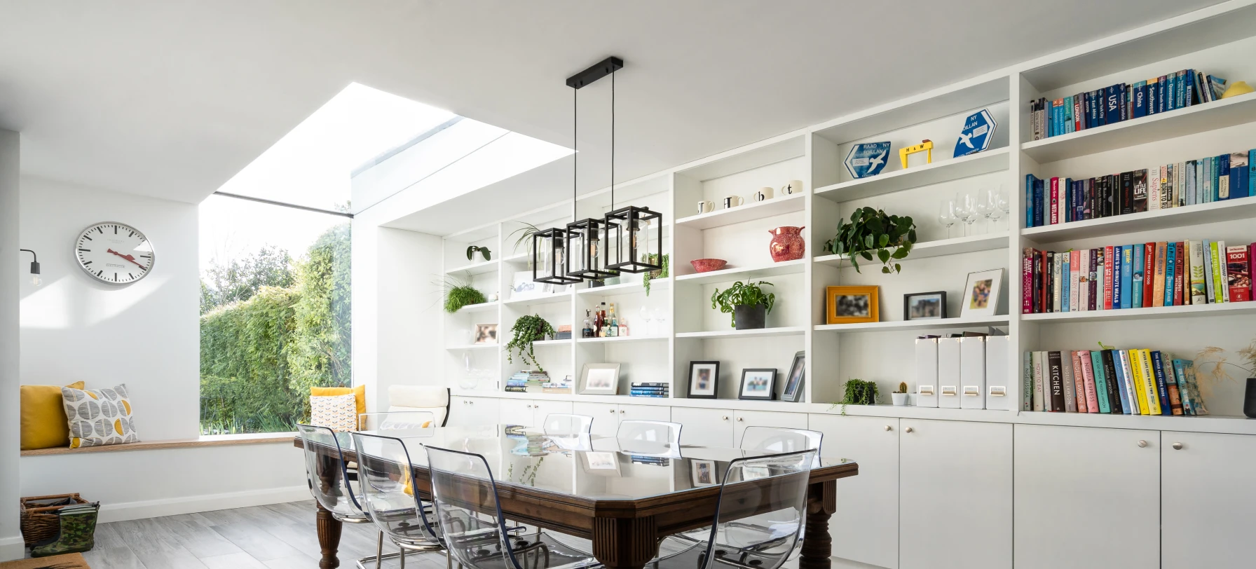 Moderner Essbereich mit einem großen Holztisch und transparenten Stühlen. Im Hintergrund befinden sich Regale mit Pflanzen und Büchern. Eine große Fensteröffnung sorgt für viel Tageslicht.
