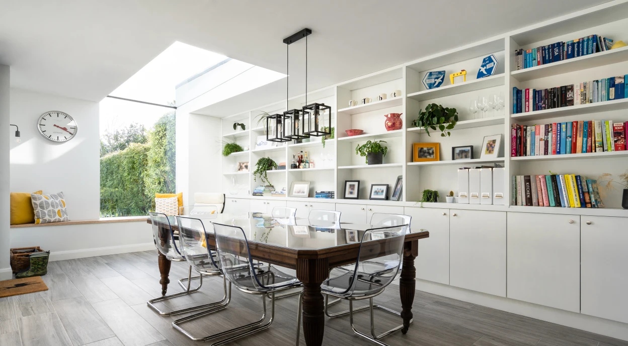 Une salle à manger moderne avec une grande table en bois et des chaises en acrylique. Un mur blanc avec des étagères remplies de livres et de plantes. Une grande fenêtre laisse entrer la lumière naturelle. Un coin salon est visible à gauche. Une horloge murale est accrochée sur le mur.