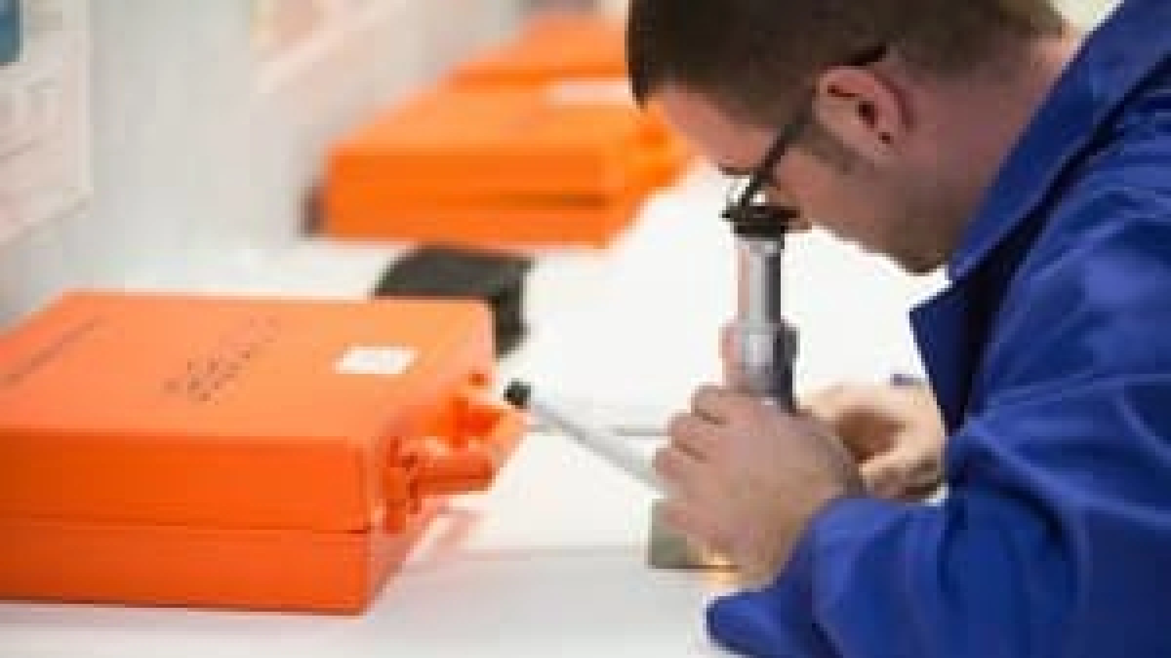 A person in a blue lab coat examines a sample under a microscope, with several orange cases visible in the background.