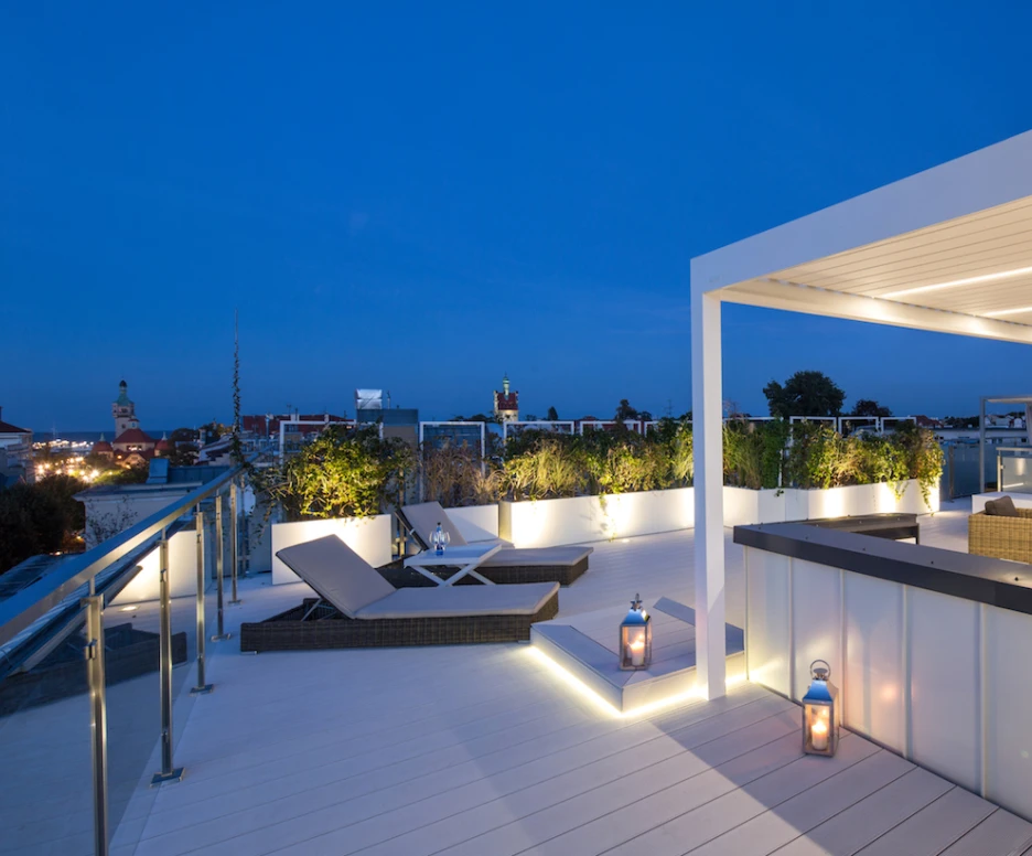 Terraza moderna al atardecer, con muebles elegantes y luces cálidas. Se aprecia una vista panorámica de la ciudad y un diseño contemporáneo con plantas decorativas. Logo de "Taras Na Fali" en la esquina inferior derecha.