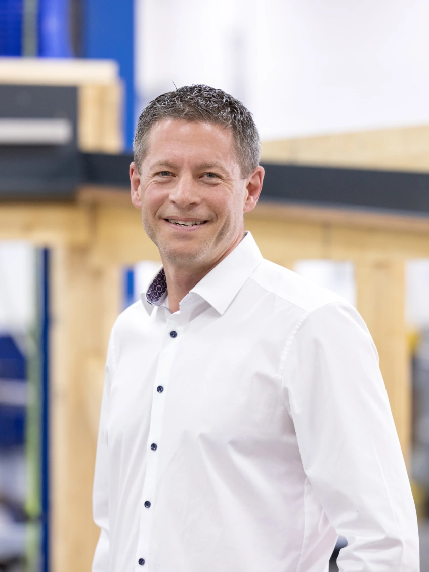 A man in a white shirt smiles confidently in a workshop setting, with wooden structures and industrial equipment visible in the background.