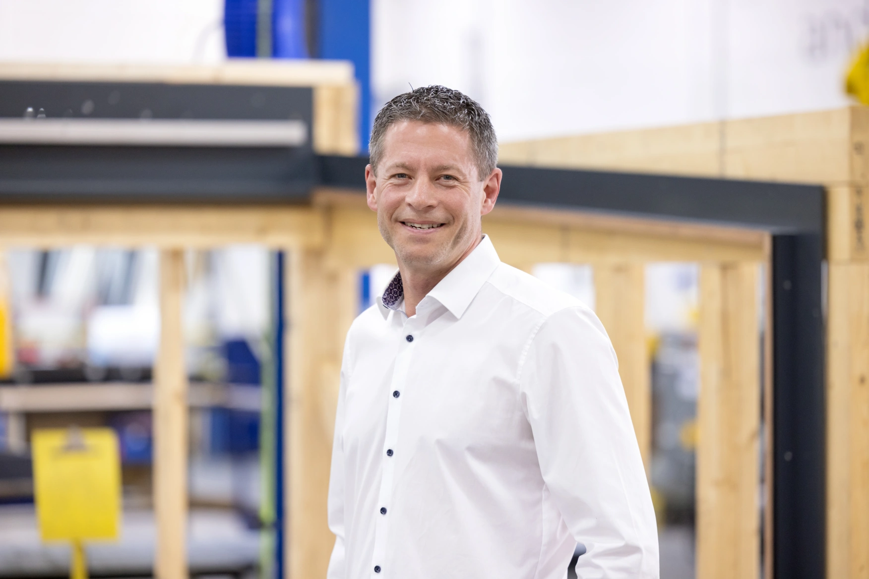 A man in a white shirt smiles confidently in a workshop setting, with wooden structures and industrial equipment visible in the background.
