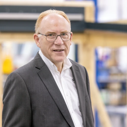 A middle-aged man with glasses wearing a dark suit and white shirt stands confidently in a workshop setting filled with wooden structures.