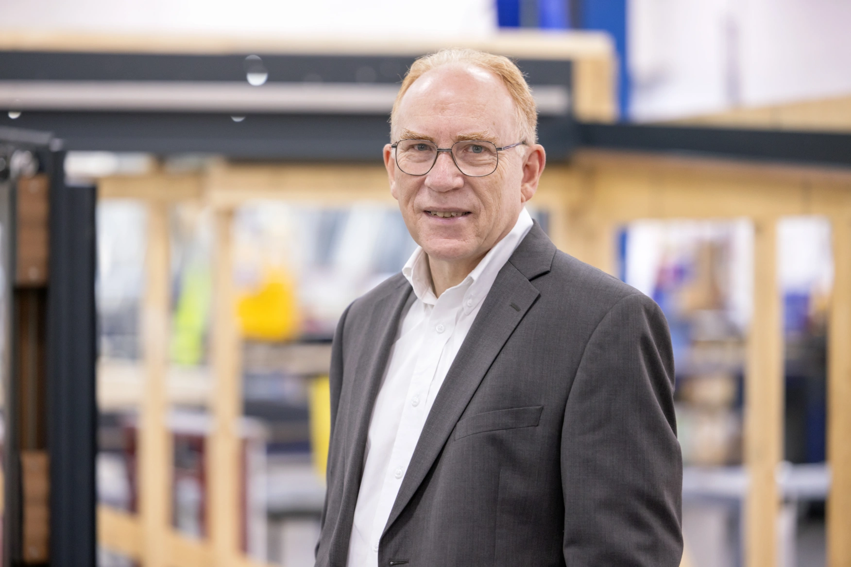 A man with glasses wearing a suit stands confidently in an industrial setting, featuring wooden structures and tools in the background.