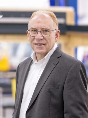 A man with glasses wearing a suit stands confidently in an industrial setting, featuring wooden structures and tools in the background.