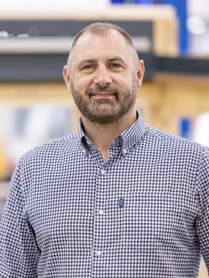 A man with a beard wearing a checkered shirt stands in a workshop with wooden shelving and tools in the background. He smiles confidently at the camera.