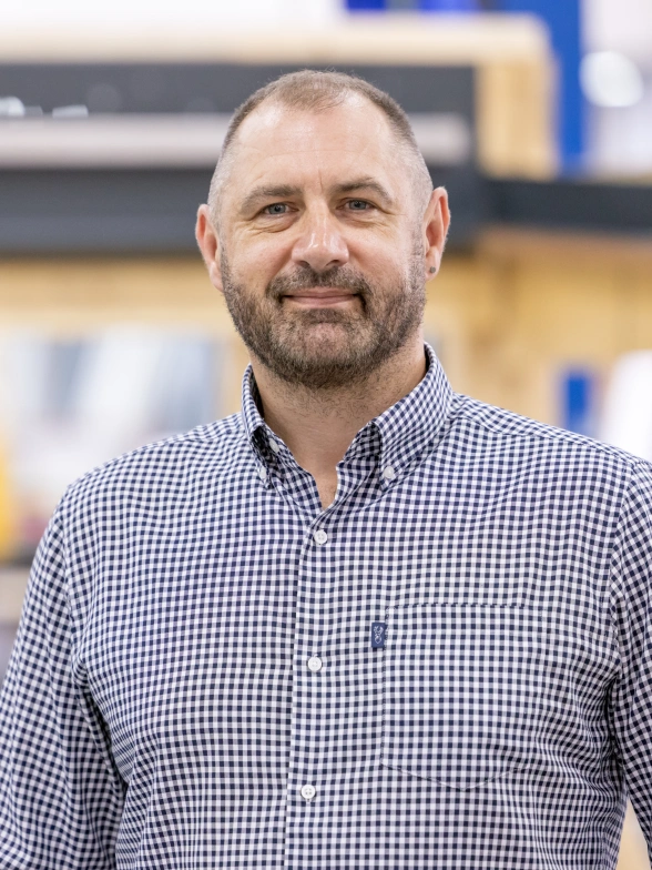 A man with a beard wearing a checkered shirt stands in a workshop with wooden shelving and tools in the background. He smiles confidently at the camera.
