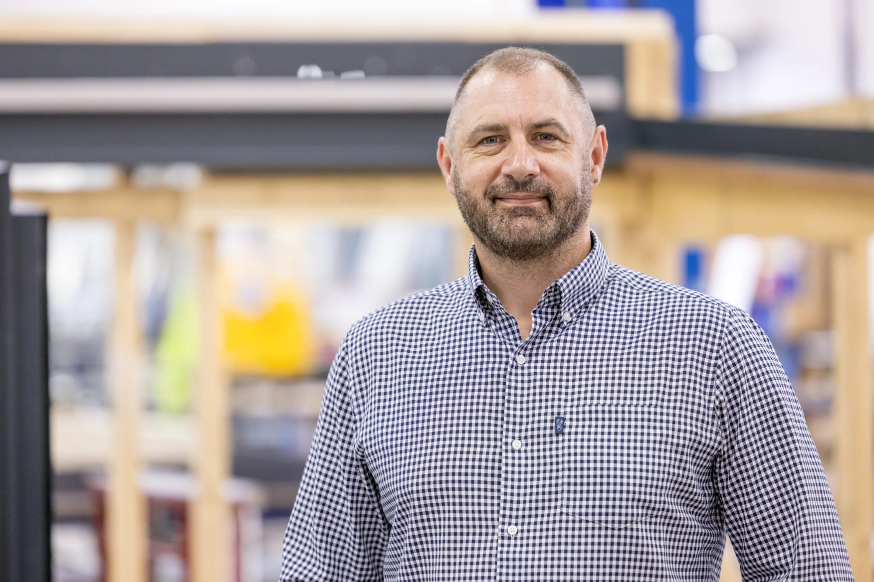 A man with a beard wearing a checkered shirt stands in a workshop with wooden shelving and tools in the background. He smiles confidently at the camera.