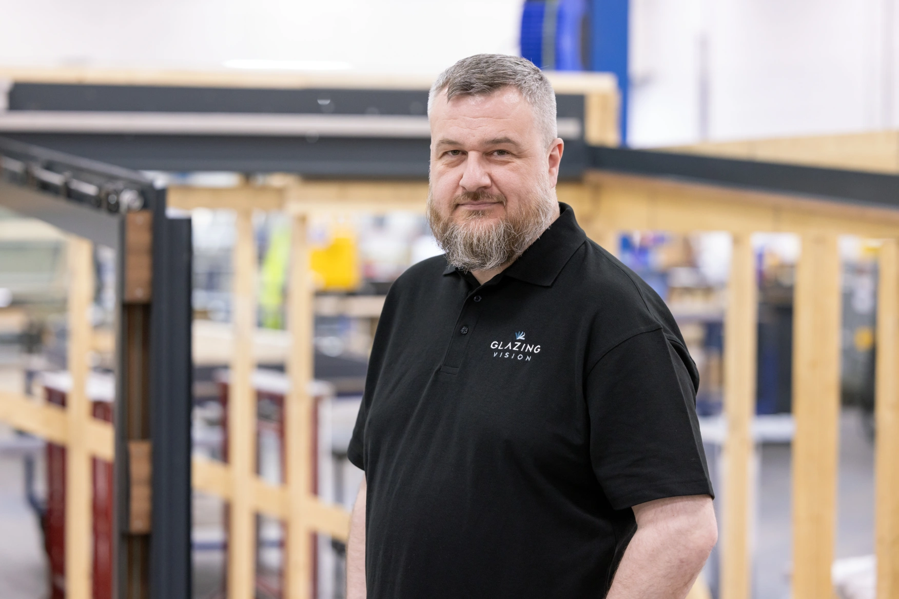 A man with a beard stands confidently in a workshop setting. He is wearing a black polo shirt with a logo that reads "Glazing Vision." Behind him, wooden frames are visible, indicating a manufacturing environment.