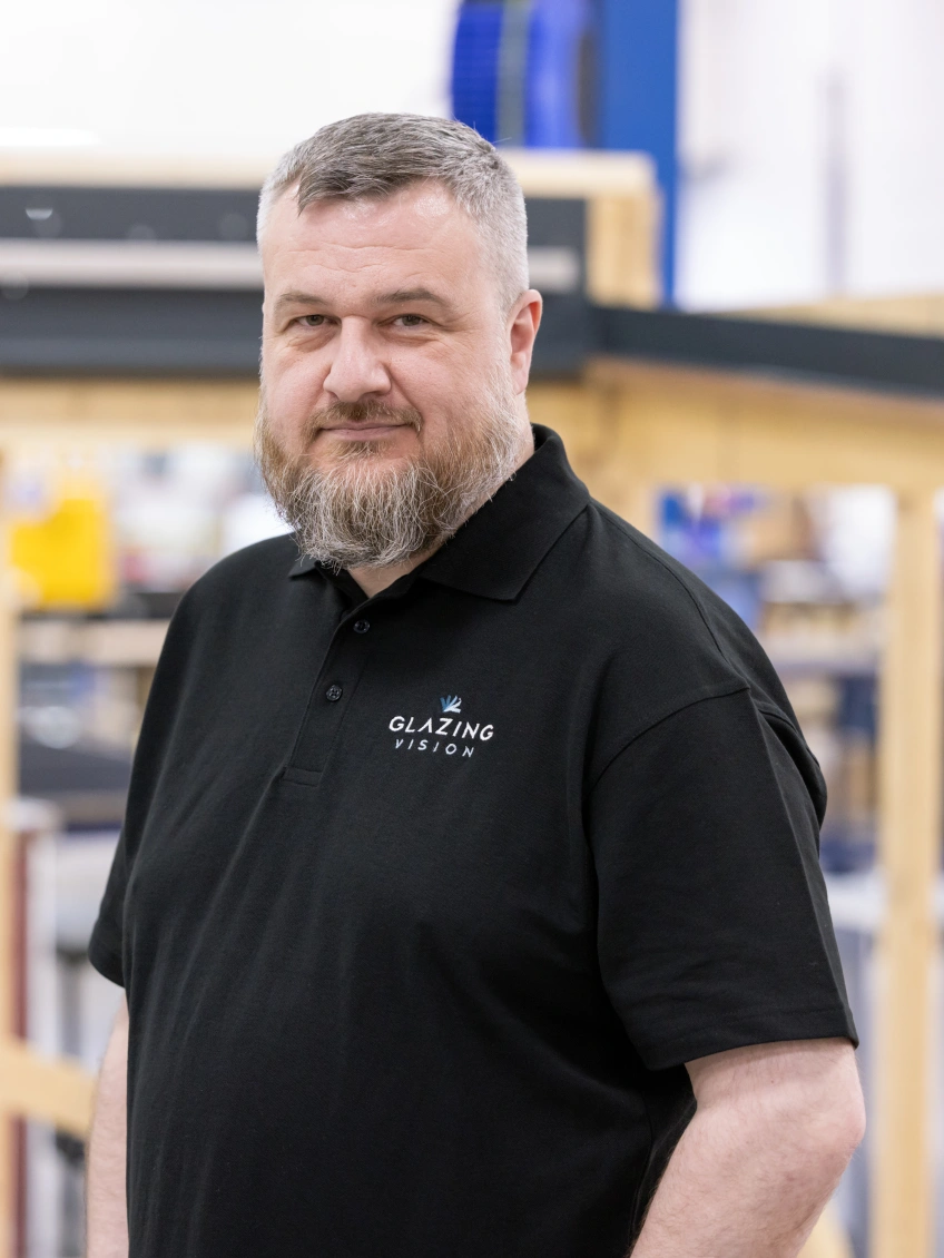 A man with a beard stands confidently in a workshop setting. He is wearing a black polo shirt with a logo that reads "Glazing Vision." Behind him, wooden frames are visible, indicating a manufacturing environment.