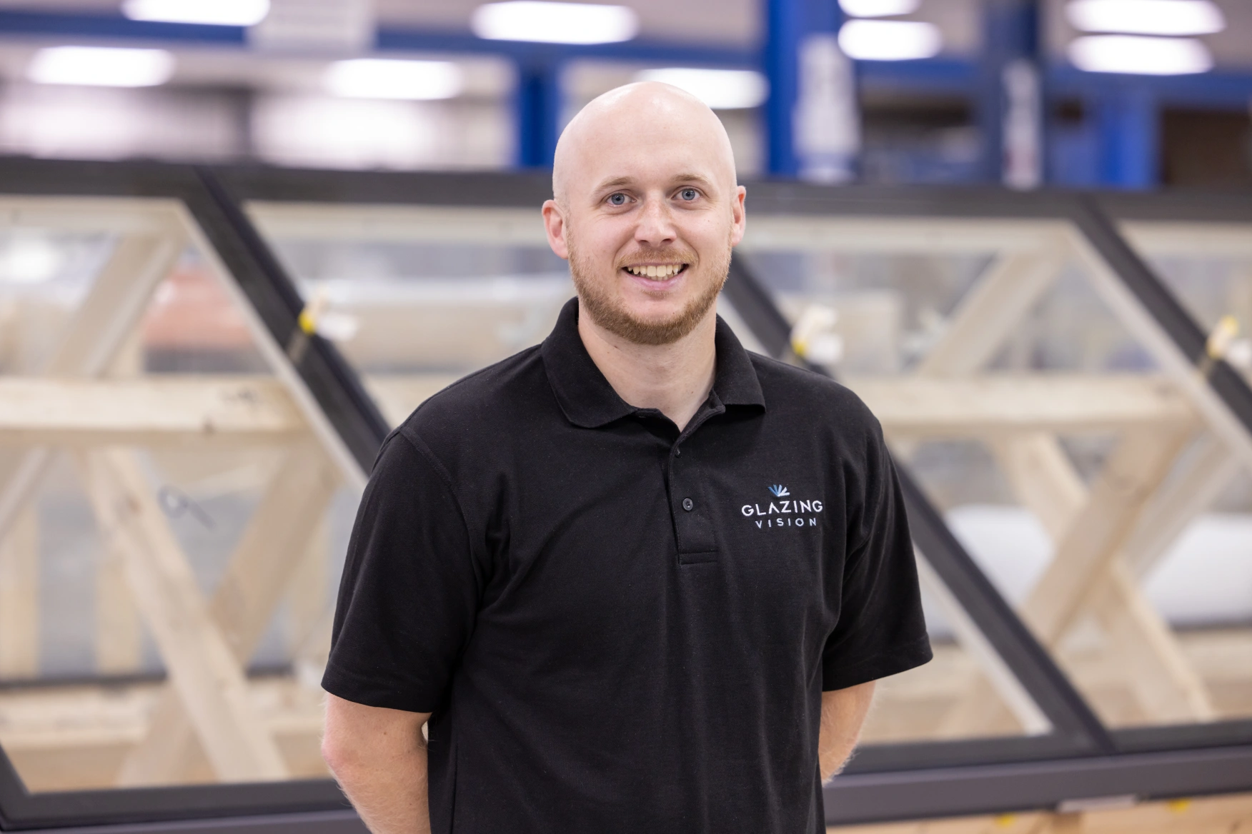 A man with a shaved head and beard smiles while standing in a warehouse. He wears a black polo shirt with the logo "Glazing Vision." In the background, there are partially assembled glass structures.