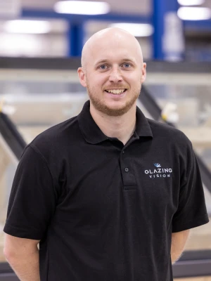 A man with a shaved head and beard smiles while standing in a warehouse. He wears a black polo shirt with the logo "Glazing Vision." In the background, there are partially assembled glass structures.
