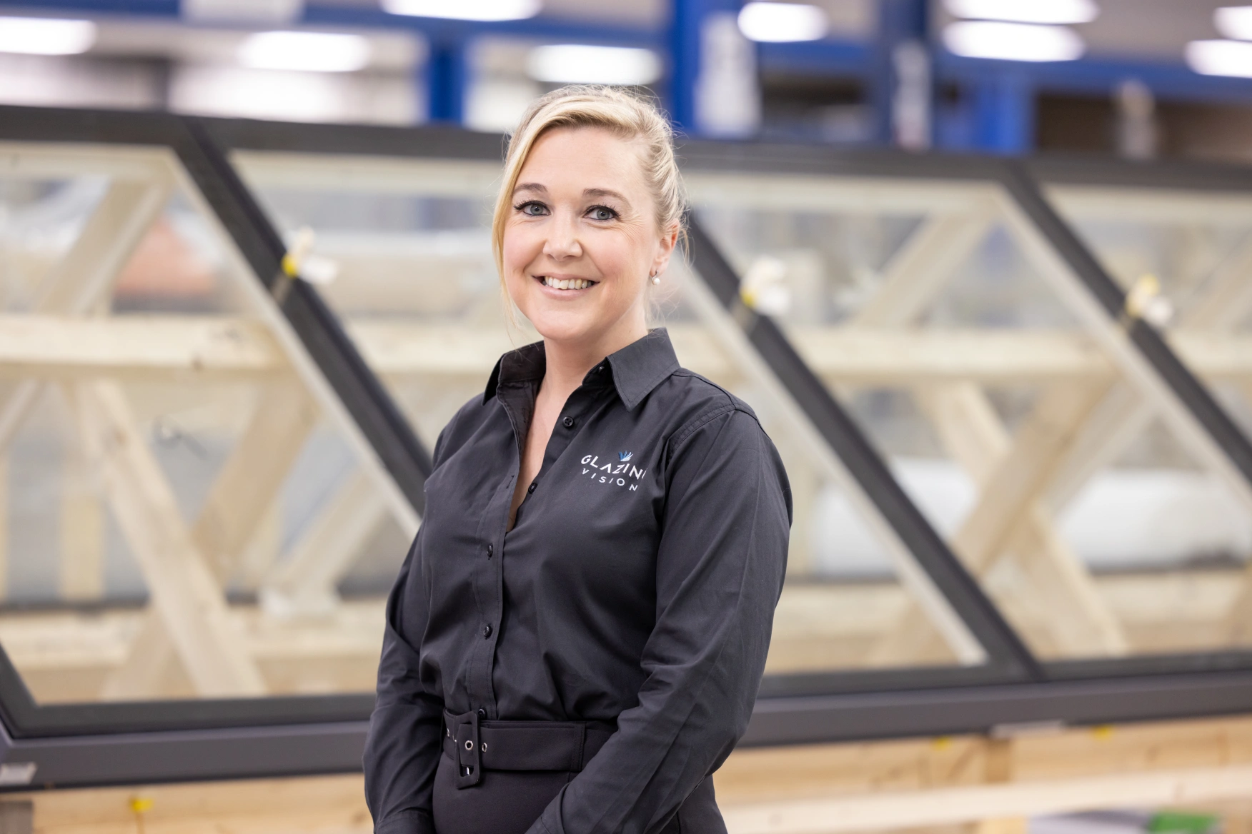 A woman in a black shirt stands smiling in front of large windows in a workshop. The background shows wooden frames and a well-lit setting.