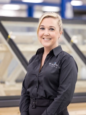 A woman with blonde hair styled in a ponytail smiles while standing in front of large windows at a manufacturing facility. She is wearing a black shirt with a logo on it. The background shows a well-lit workspace filled with window frames.