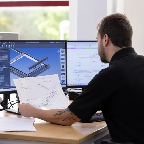 Ein Mann sitzt an einem Schreibtisch vor zwei Computermonitoren und betrachtet einen Plan. Auf dem Bildschirm ist eine technische Zeichnung zu sehen. Im Hintergrund sind Fenster mit Tageslicht.