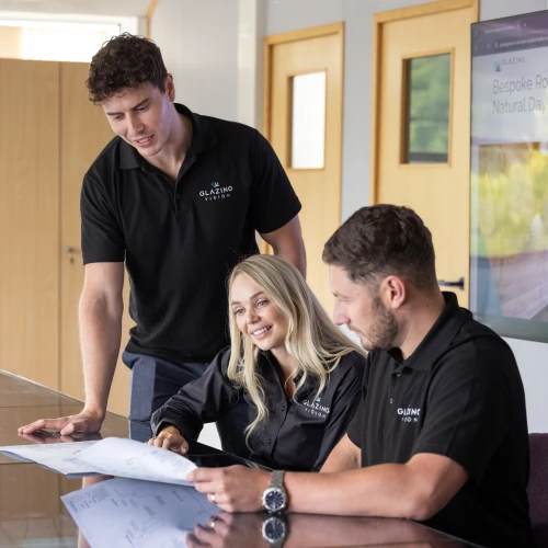 Three people collaborating on a project at a table. A man stands to the left, while a woman and another man sit at the table, reviewing documents. All three are wearing black polo shirts with a logo. A screen in the background displays information.