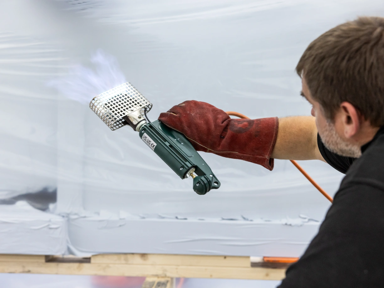 A person wearing red gloves uses a spray gun to apply a coating on a surface. The background is covered with a plastic sheet, and there are visible blue spray marks.