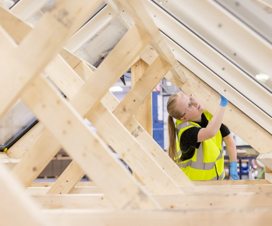 Eine Person mit einer Warnweste und Schutzbrille arbeitet an einer Holzstruktur, die Teil eines Bauprojekts ist. Sie prüft sorgfältig die Konstruktion unter einem Dachfenster.