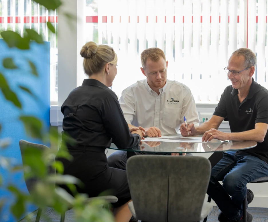 Three people seated at a round table in a bright office. One person is taking notes while the others discuss a document. Green plant leaves are visible in the foreground.