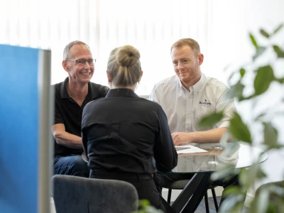 Two men and a woman are sitting around a table in a bright office. The woman, facing away from the camera, is dressed in a black shirt, while the men are engaged in conversation, smiling. One man is wearing glasses and a black shirt, and the other is in a white shirt with a logo. Green plants are visible in the foreground.