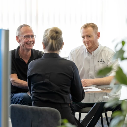 Two men are sitting at a glass table, engaging in a discussion with a woman who has her back to the camera. The setting features bright indoor lighting and plants in the background. The men appear to be smiling and interacting positively.