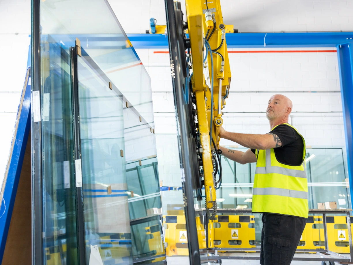 A worker in a safety vest operates a glass handling machine to lift large panes of glass in a manufacturing facility. The environment is spacious with metal structures and safety barriers visible in the background.