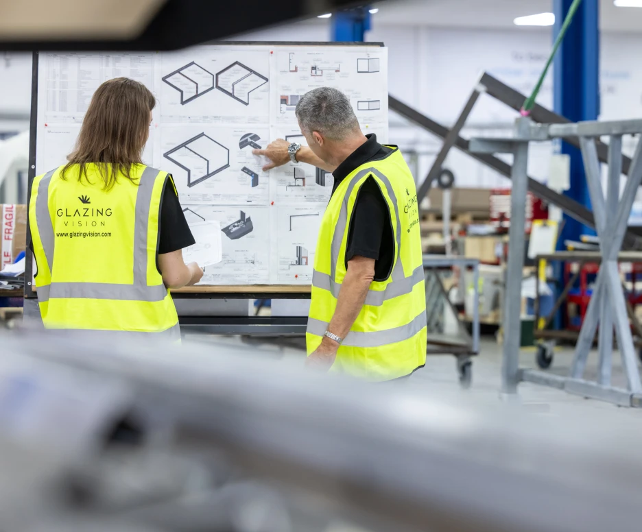 Two individuals in high-visibility vests examine a blueprint displayed on an easel in a workshop. One person points at a specific section of the plan while the other observes closely. In the background, industrial equipment and tools are visible.