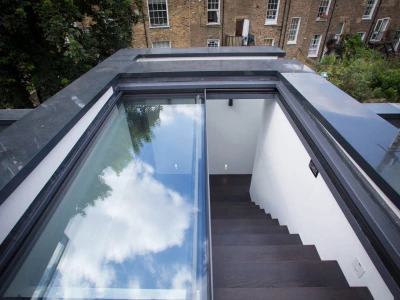 A modern staircase leads down from a rooftop terrace, featuring glass panels on either side. The scene includes clear blue skies and clouds reflected in the glass above, with brick buildings visible in the background.