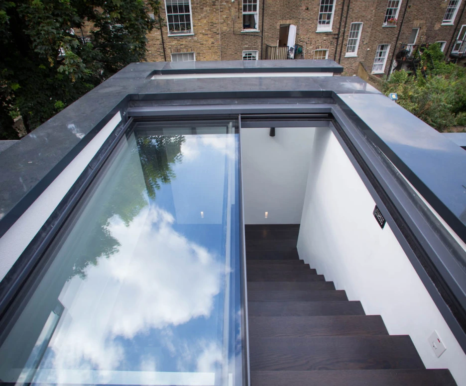 A modern staircase leads down from a rooftop terrace, featuring glass panels on either side. The scene includes clear blue skies and clouds reflected in the glass above, with brick buildings visible in the background.