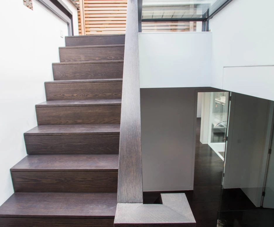 A modern interior staircase with dark wooden steps and a sleek railing, leading to a bright upper level. The walls are clean and minimalistic, with glass elements adding a contemporary touch.