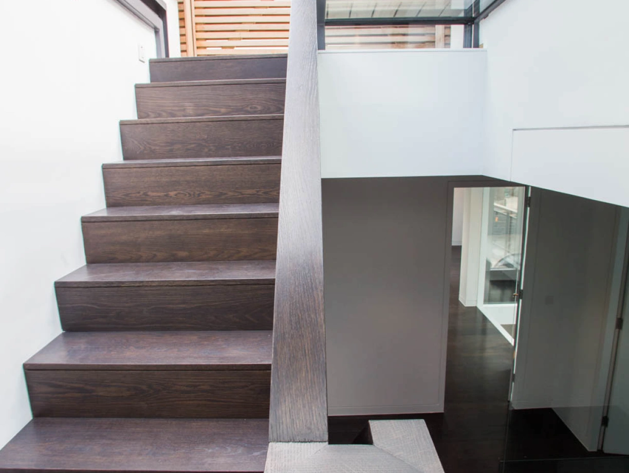 A modern interior staircase with dark wooden steps and a sleek railing, leading to a bright upper level. The walls are clean and minimalistic, with glass elements adding a contemporary touch.