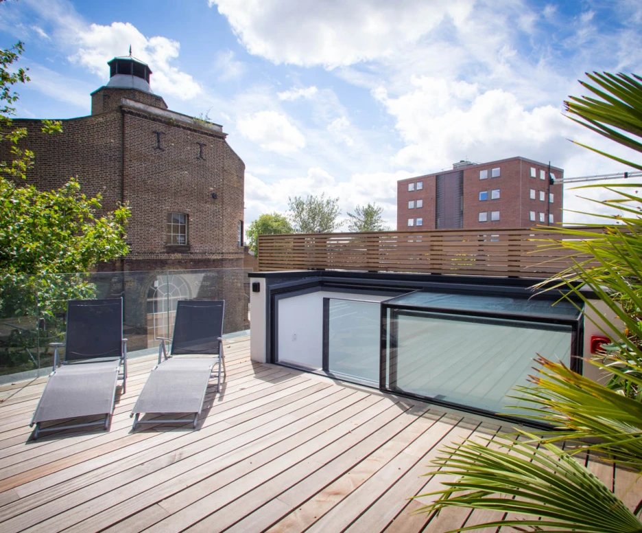 A rooftop terrace features two lounge chairs on wooden decking, surrounded by greenery. In the background, a historic brick building and a modern apartment complex are visible under a partly cloudy sky.