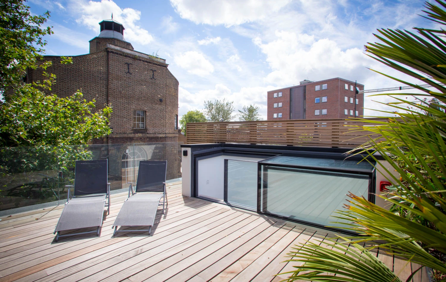A spacious wooden deck features two lounge chairs under a bright sky with scattered clouds. In the background, a historic brick building and a modern apartment building are visible. Lush green foliage surrounds the area, creating a relaxing atmosphere.