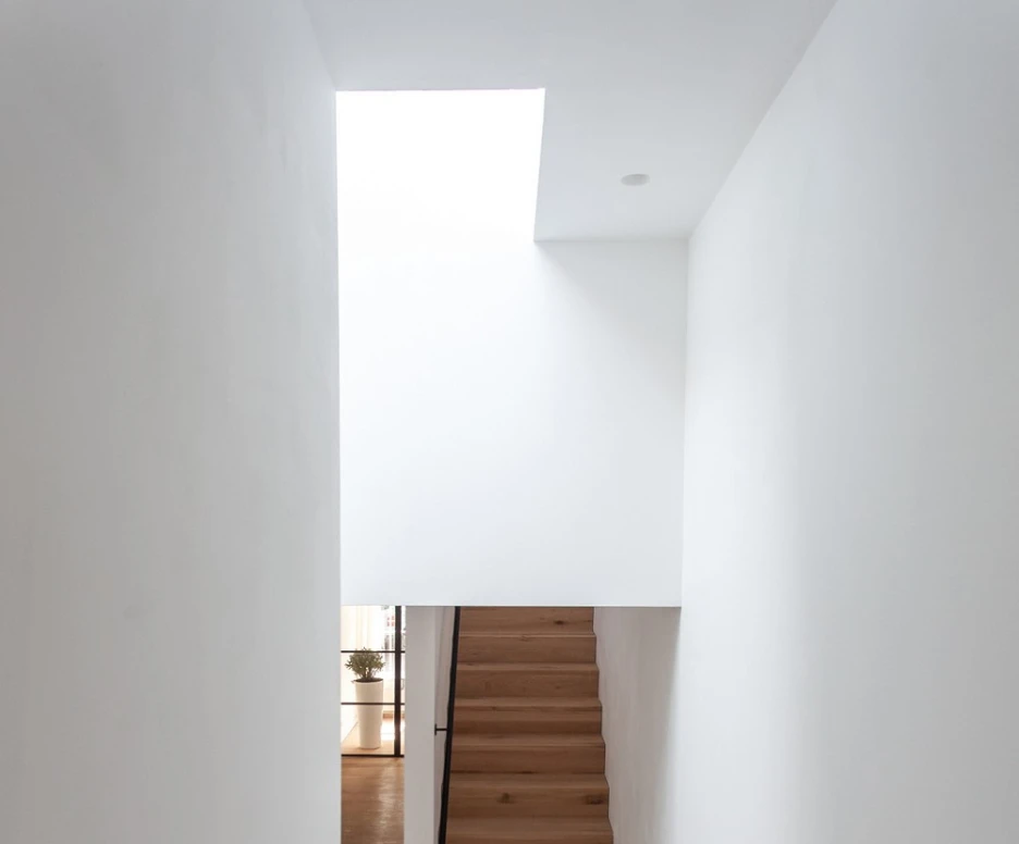 A wooden staircase ascends in a light-filled hallway, featuring a skylight above. The walls are painted white, creating a bright and minimalistic atmosphere.