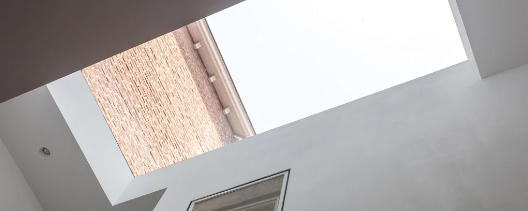 View of a modern interior with a skylight. The ceiling is white, and the skylight frames the sky above, contrasting with the brick building visible through the opening.
