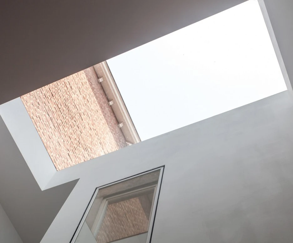 View of a modern interior with a skylight. The ceiling is white, and the skylight frames the sky above, contrasting with the brick building visible through the opening.