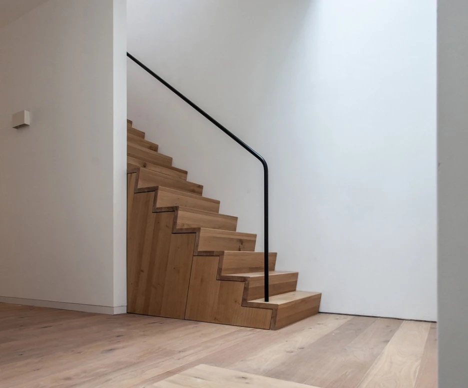 A modern staircase with wooden steps and a black handrail, leading upwards in a minimalist space. Natural light streams in from a skylight above, illuminating the clean white walls and wooden flooring.