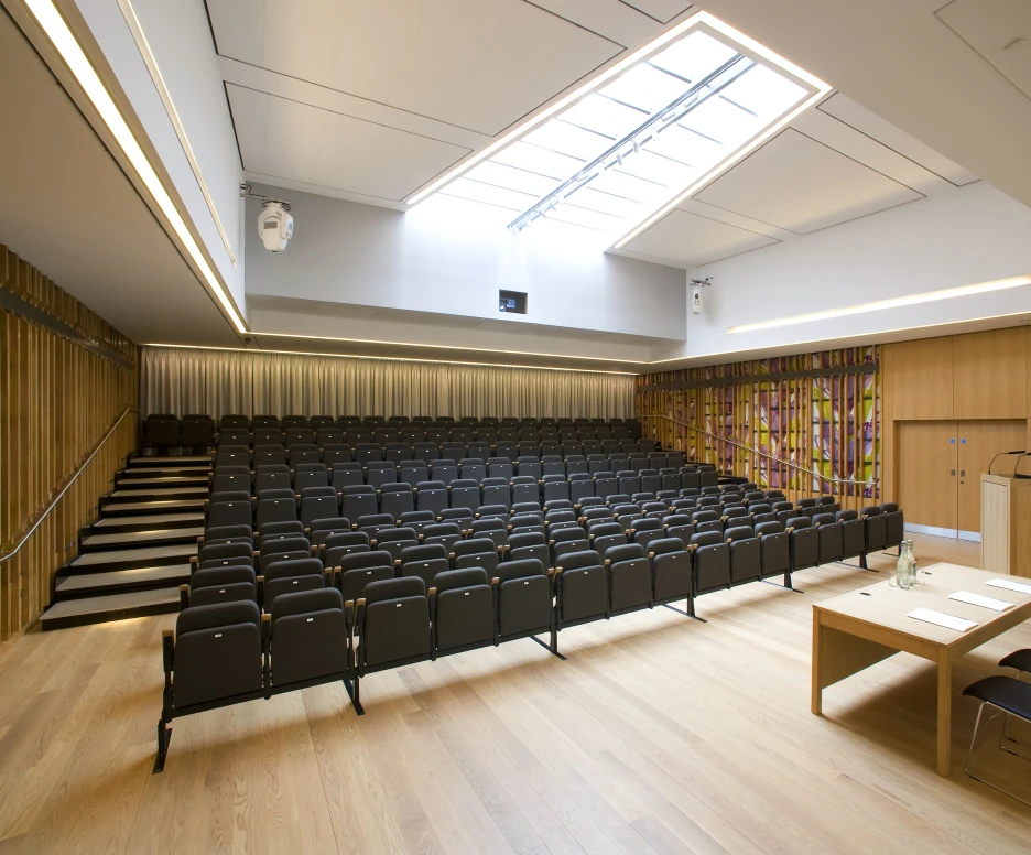 Salle de conférence moderne avec des rangées de chaises noires, un podium en bois et des fenêtres au plafond. Les murs en bois et un motif coloré ajoutent une touche contemporaine.