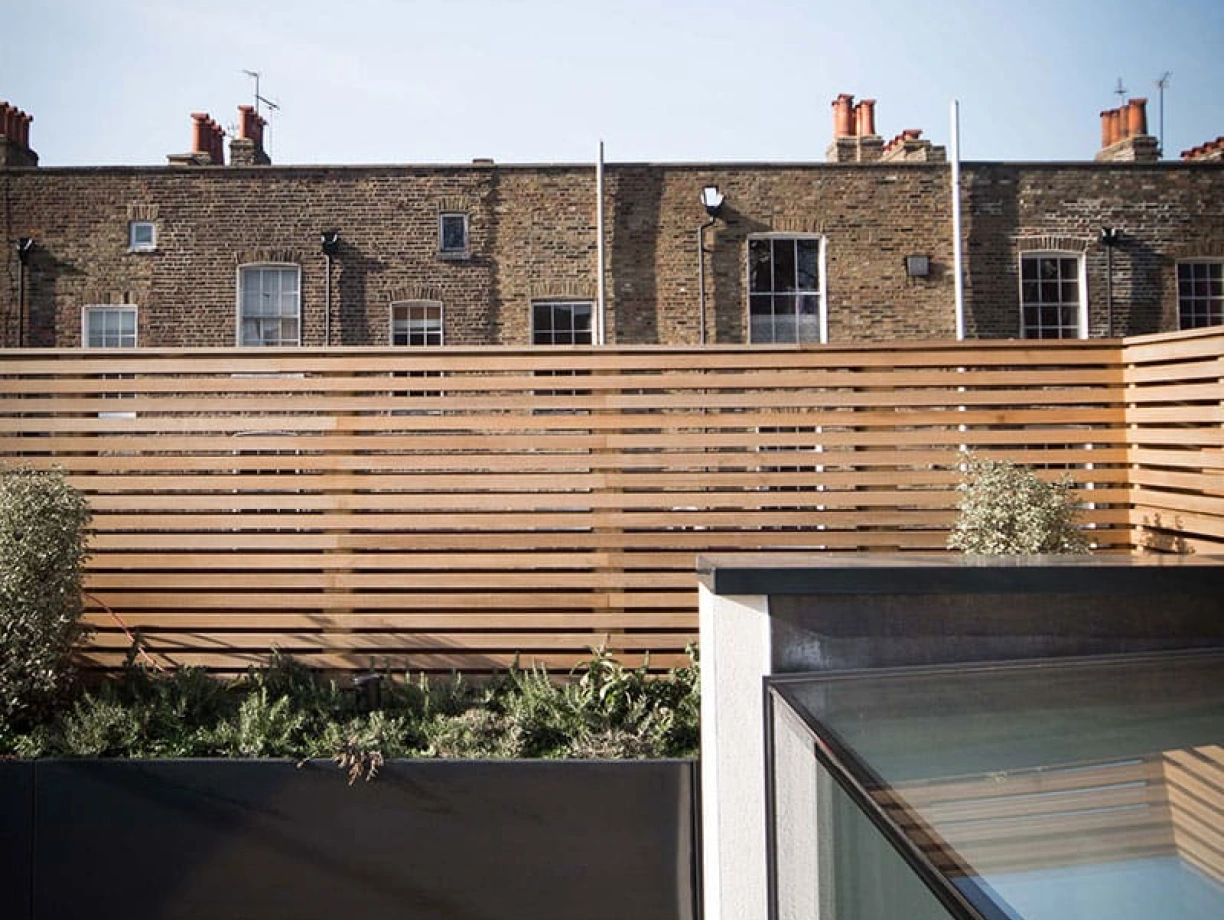 A wooden deck area with a black railing overlooks a building with brick walls and red chimneys in the background. Vertical wooden slats create a privacy barrier on one side. The sky above is clear and blue.