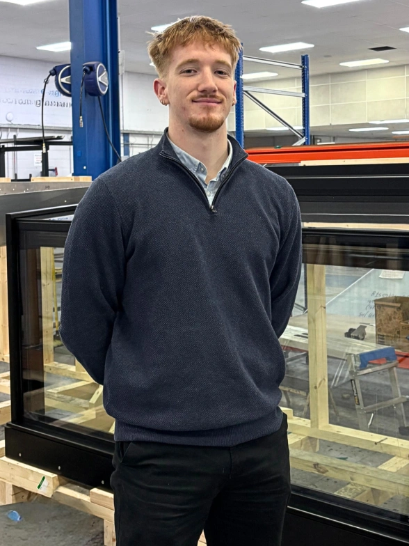 A young man stands confidently in a warehouse setting, wearing a dark sweater and black pants. He has short, light brown hair and is smiling slightly. In the background, there are wooden structures and equipment, indicating a working environment.