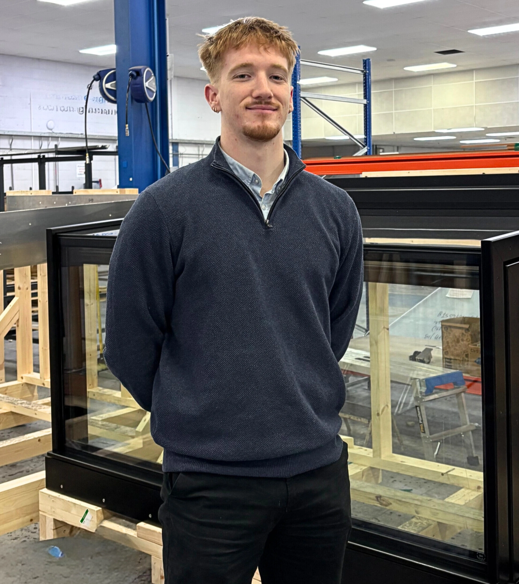A young man stands confidently in a warehouse setting, wearing a dark sweater and black pants. He has short, light brown hair and is smiling slightly. In the background, there are wooden structures and equipment, indicating a working environment.