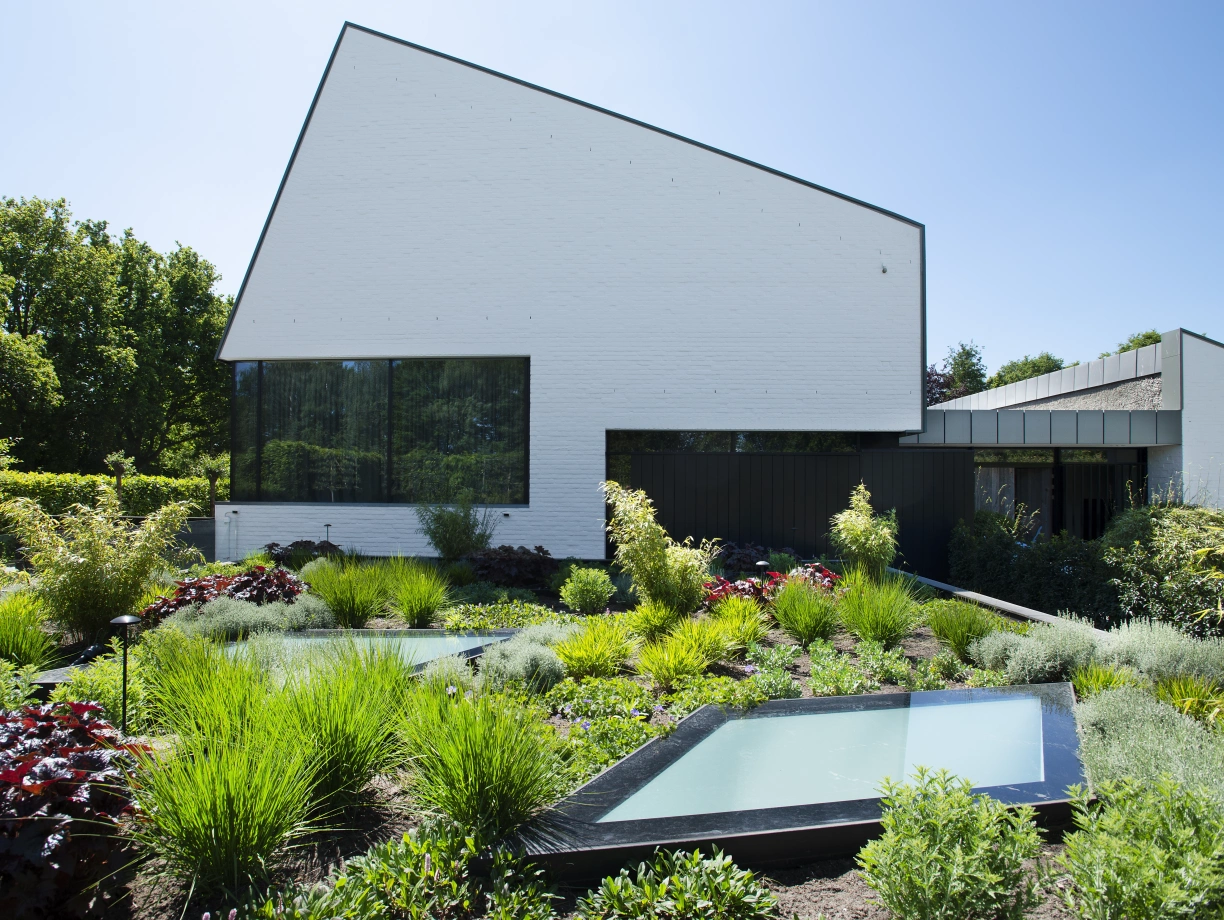 A modern white house with a distinctive angular roof is surrounded by lush greenery and landscaped gardens. Two shallow water features are integrated into the garden, reflecting the vibrant foliage. Bright blue sky is visible above, enhancing the serene outdoor atmosphere.